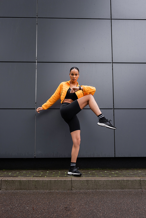 full length of stylish african american sportswoman in bike shorts and yellow puffer jacket exercising outside