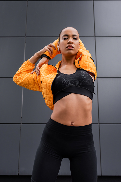 low angle view of african american sportswoman in sports bra and yellow puffer jacket warming up near grey building
