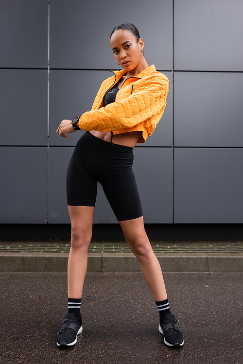 full length of stylish african american sportswoman in bike shorts and yellow puffer jacket posing near grey building