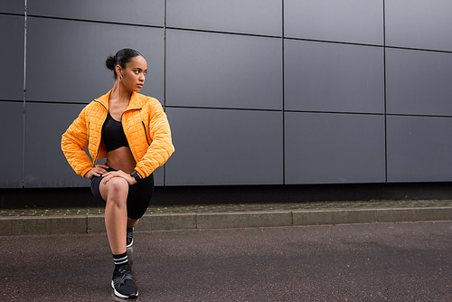 full length of stylish african american sportswoman in bike shorts and yellow puffer jacket working out near grey building