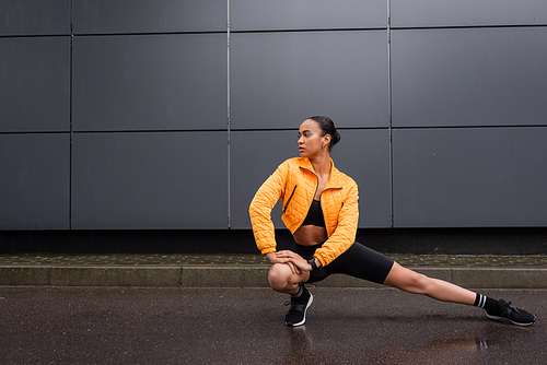 full length of stylish african american sportswoman in bike shorts and yellow puffer jacket stretching near grey building