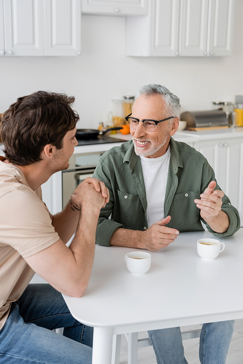cheerful and mature father in eyeglasses gesturing while talking with son near cups of coffee