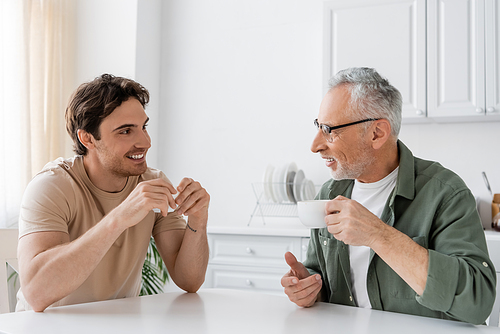 happy young man drinking morning coffee with smiling and mature father at home