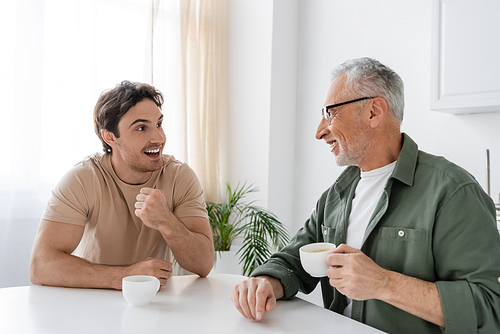 excited guy gesturing while talking to smiling dad holding cup of coffee in kitchen