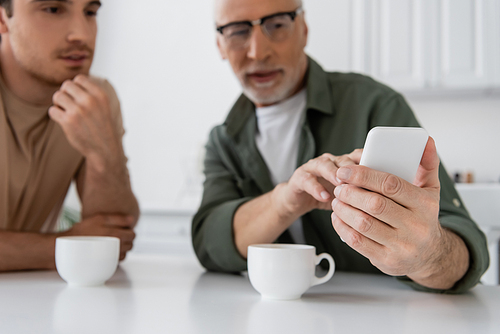 mature man in eyeglasses pointing at smartphone near adult son and coffee cups on blurred background