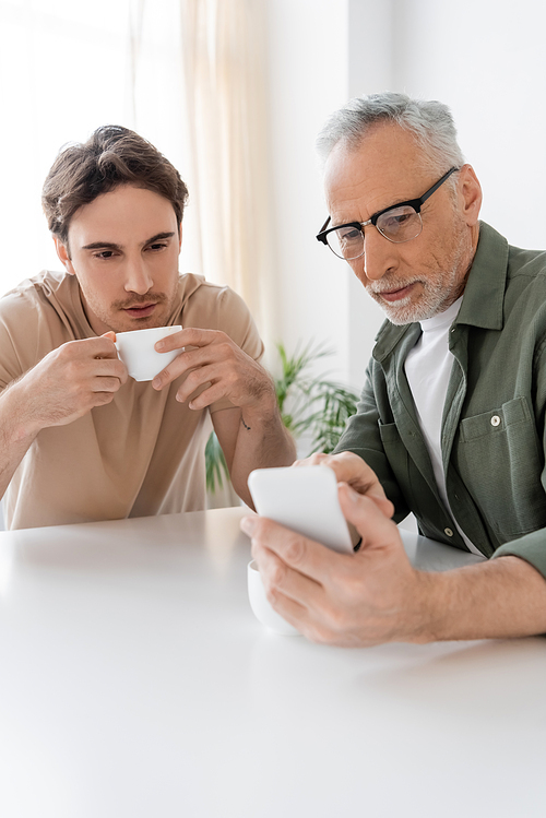 senior man in eyeglasses showing smartphone to young son drinking coffee in kitchen