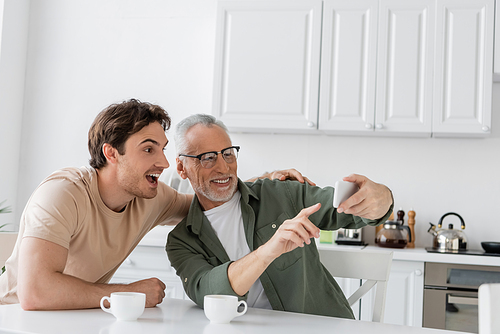 smiling grey haired man pointing at mobile phone near amazed son and coffee cups on kitchen table