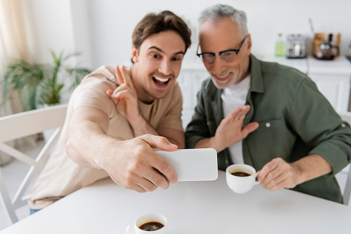 cheerful young man showing victory sign while taking selfie with mature dad holding coffee cup and waving hand