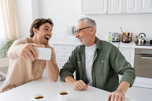 young excited man with mobile phone taking selfie with smiling father near coffee cups in kitchen