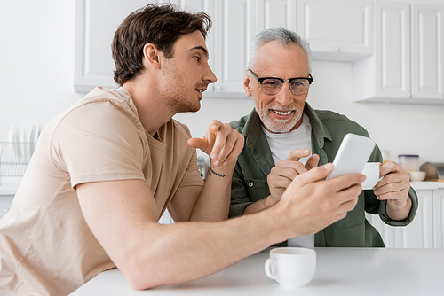 young man showing mobile phone to cheerful father sitting with coffee cup in kitchen