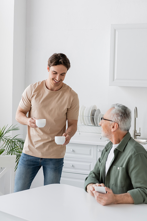young man smiling while holding coffee cups near dad sitting with smartphone in kitchen