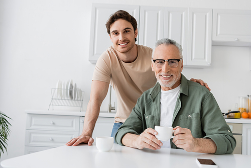 happy mature man with coffee cup and happy young son looking at camera in kitchen