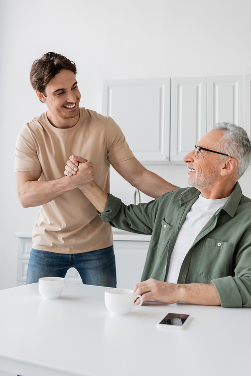 happy dad and son shaking hands near coffee cups and mobile phone with blank screen on kitchen table