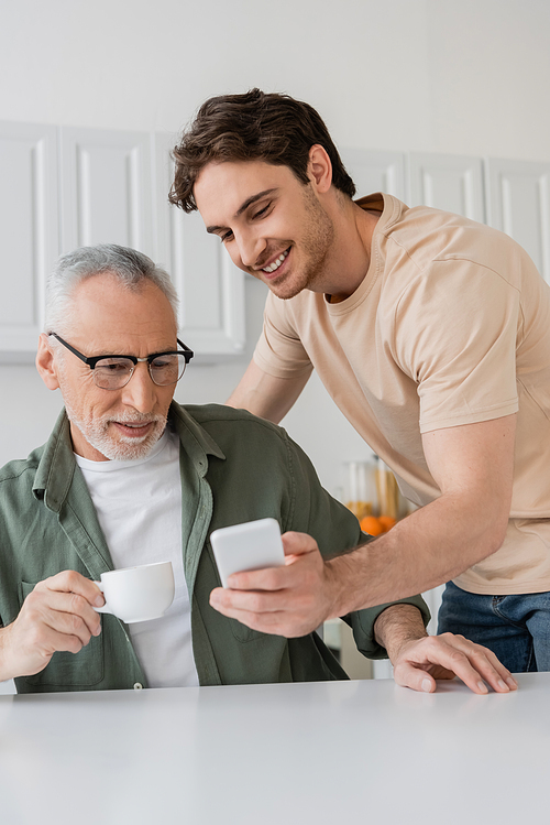 young man showing mobile phone to father drinking morning coffee in kitchen