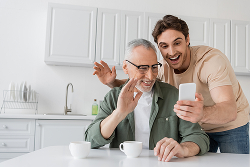 cheerful man with adult son waving hands during video call on mobile phone in kitchen