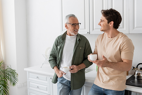 smiling mature man holding coffee cup while standing with hand in pocket and listening to son in kitchen