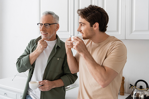 young man with coffee cup looking at thoughtful and smiling father in kitchen