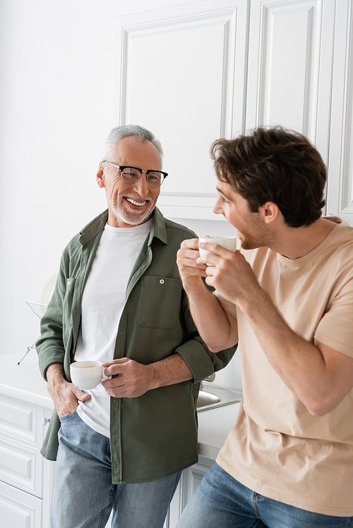 young man holding coffee cup and looking at cheerful dad during conversation in kitchen