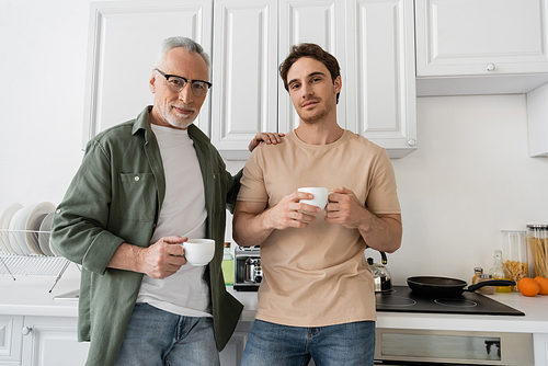 mature man in eyeglasses and young son holding coffee cups and looking at camera in kitchen