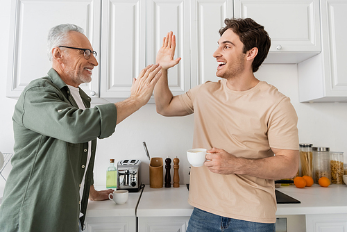 smiling man giving high five to excited son with coffee cup in kitchen