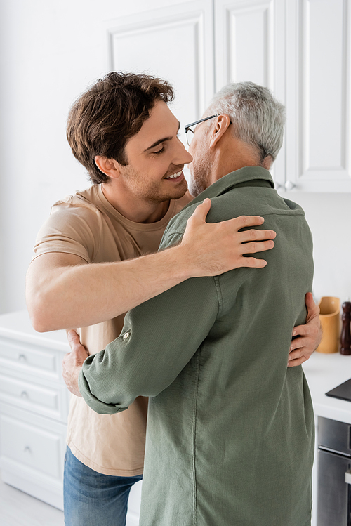 young and joyful man embracing grey haired father at home in kitchen