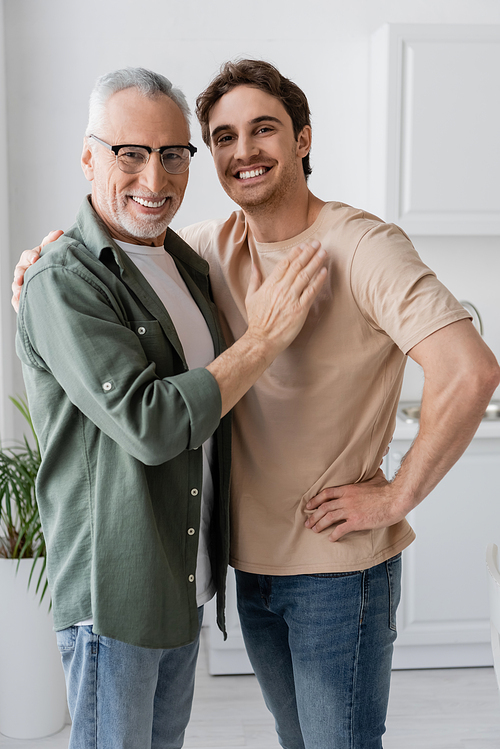 young and smiling man hugging shoulders of happy father touching his chest and smiling at camera