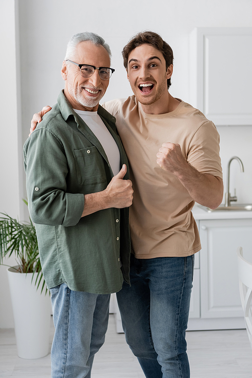 cheerful father and son showing like and success gestures while looking at camera in kitchen
