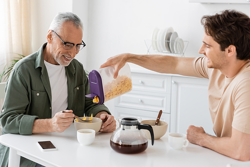young man pouring corn flakes into bowl near happy dad and coffee pot in kitchen