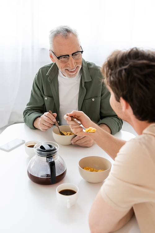 smiling man in eyeglasses looking at young son eating corn flakes for breakfast
