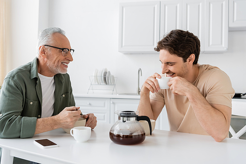 smiling man looking at son holding coffee cup and laughing with closed eyes during breakfast