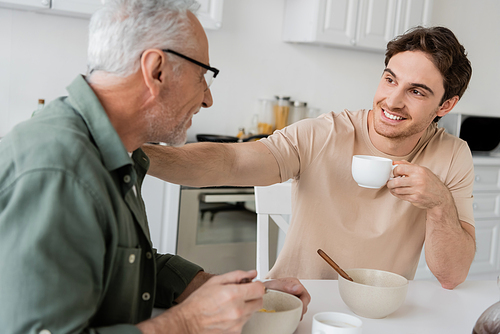 cheerful young man holding coffee cup and touching shoulder of dad while talking to him during breakfast