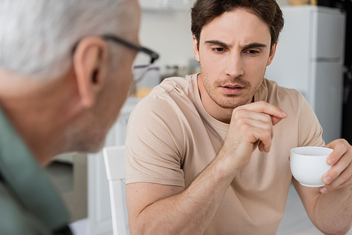 thoughtful and serious man holding coffee cup while talking to blurred father in kitchen