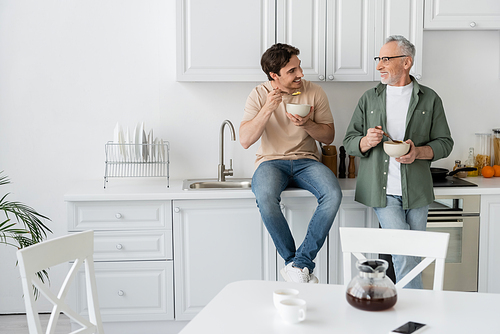 young man sitting on kitchen worktop while having breakfast and talking to smiling dad