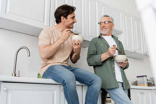 smiling guy sitting on kitchen worktop and looking at happy dad during breakfast