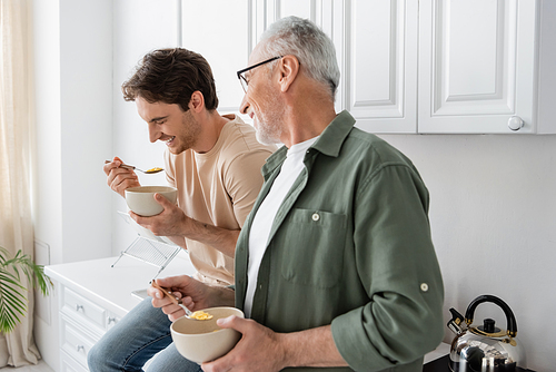 grey haired man looking at laughing son sitting on kitchen worktop with spoon and bowl of corn flakes