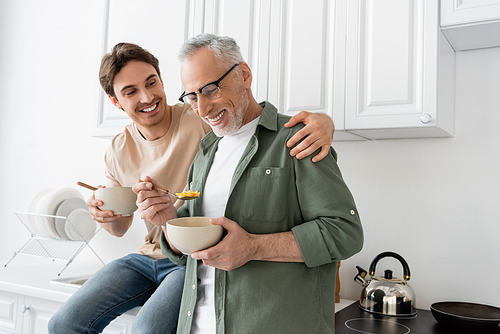 happy young man sitting on kitchen worktop and hugging shoulders of smiling dad holding bowl and spoon with corn flakes