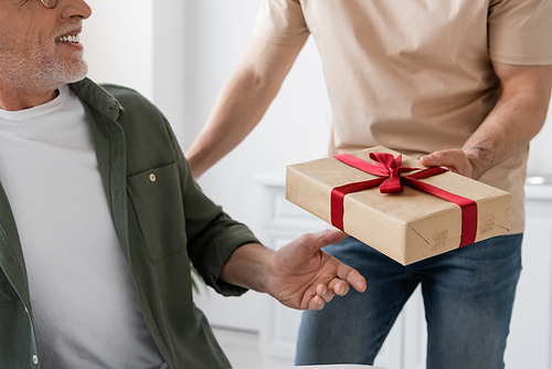 cropped view of young man holding gift box with red ribbon near smiling dad