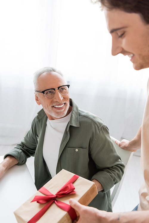 cheerful man in eyeglasses looking at blurred son holding fathers day present