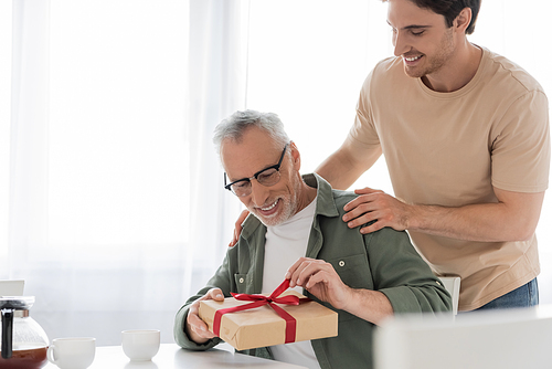 smiling guy touching shoulders of pleased dad opening fathers day present