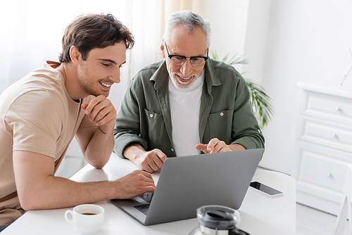 happy mature man with smiling son looking at laptop near coffee pot in kitchen