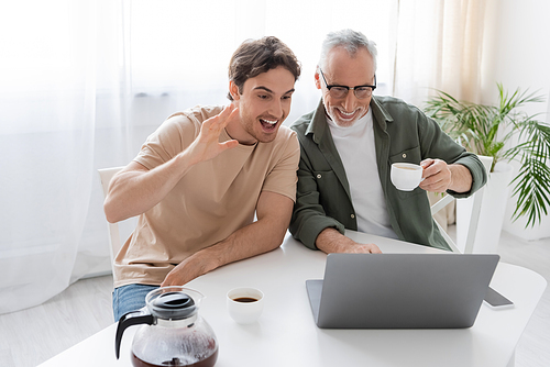 excited man waving hand during video call on laptop near cheerful father with coffee cup