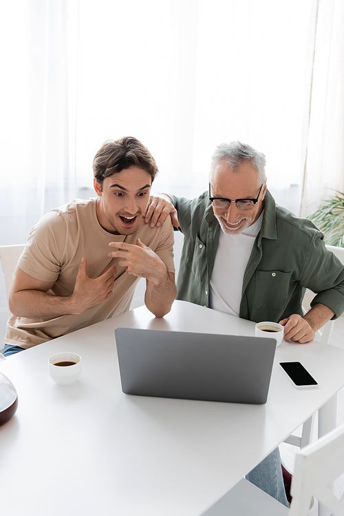surprised guy pointing at himself during video chat on laptop near mature dad and coffee cups