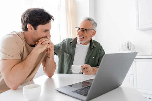 cheerful grey haired man with coffee cup looking at young son smiling with closed eyes near laptop
