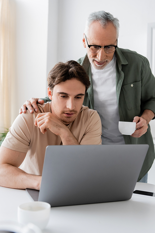 senior man with coffee cup touching shoulder of thoughtful son looking at laptop in kitchen