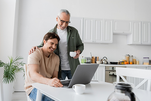 mature and smiling man with coffee cup touching shoulder of son laughing near laptop in kitchen