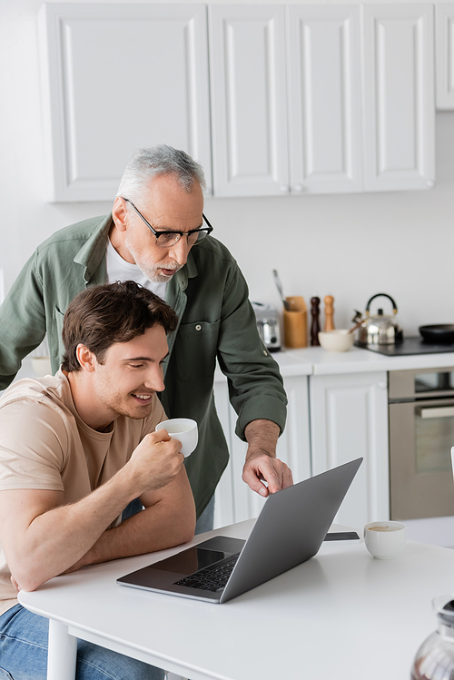 surprised man in eyeglasses pointing at laptop near smiling son drinking morning coffee in kitchen