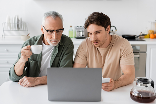 mature man with coffee cup and his young son with smartphone looking at laptop in kitchen
