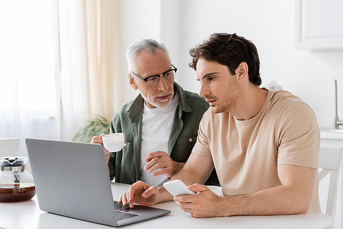 grey haired man in eyeglasses holding coffee cup and pointing at laptop near adult son