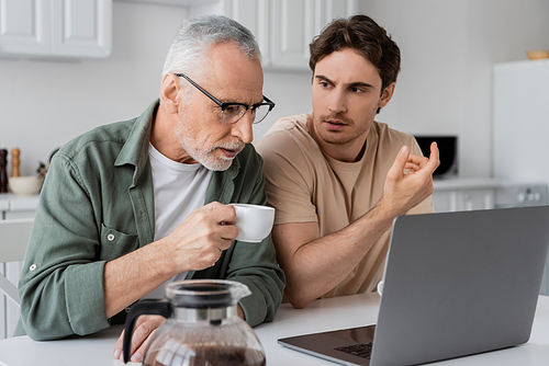 young man gesturing while talking to mature dad drinking coffee and looking at laptop in kitchen