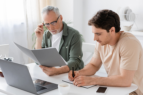thoughtful senior man in eyeglasses looking at documents near laptop and son writing in notebook